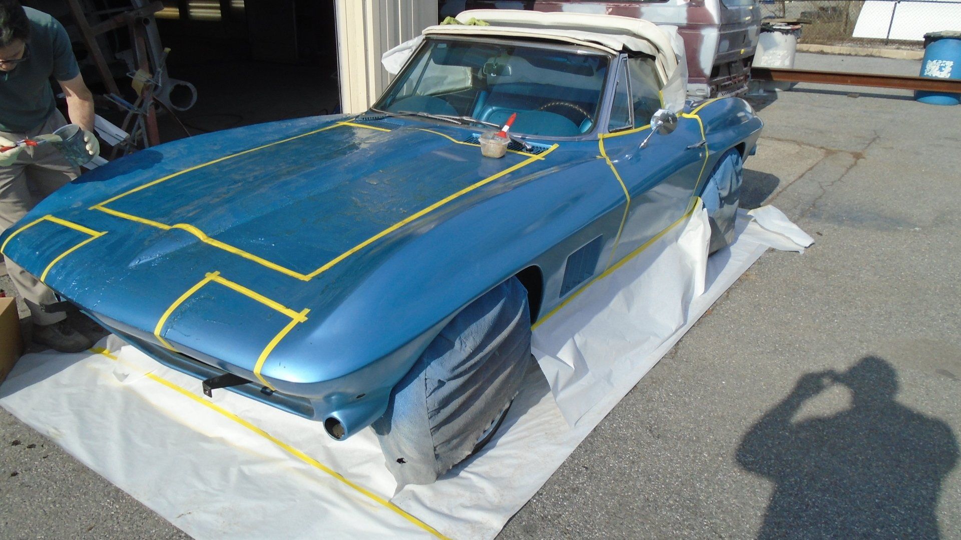 Blue classic convertible car being prepped for painting, masked with yellow tape.