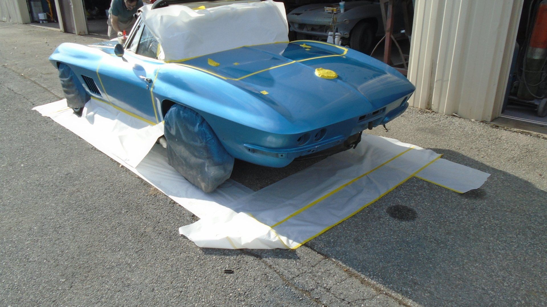 Blue convertible car covered in white protective material, taped to a garage floor for painting.