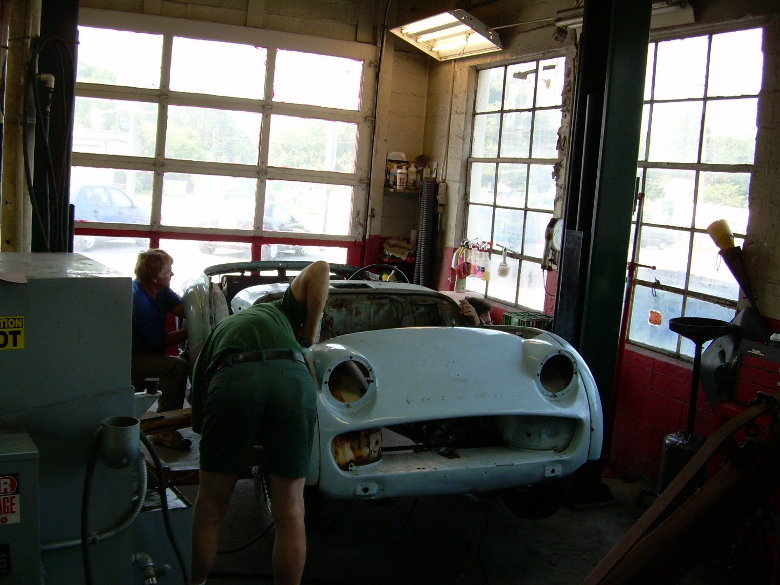 Person working on a light-blue sports car inside a garage with large windows; another person visible in the background.