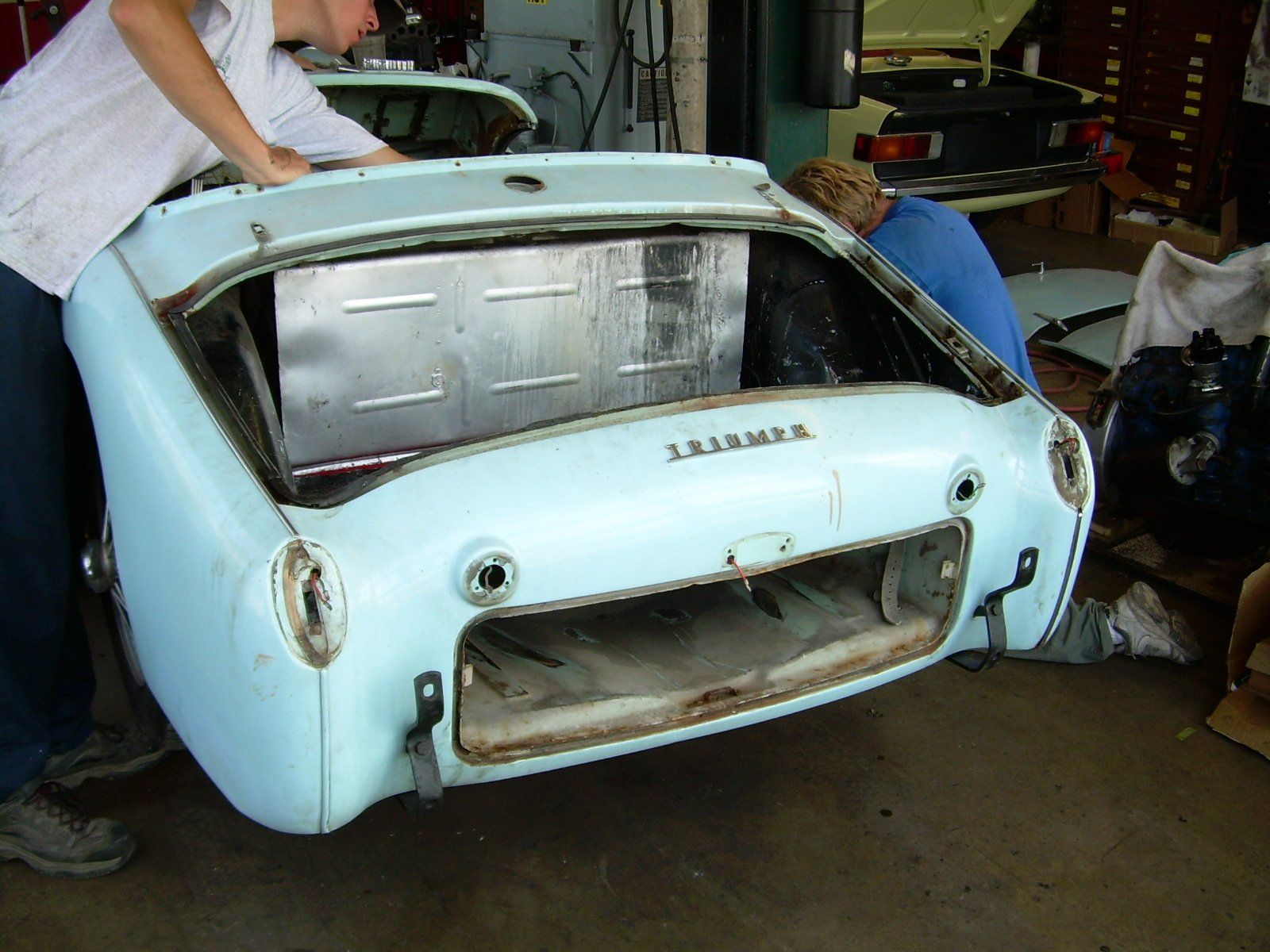 Two people working on a light blue Triumph sports car's rear section, in a workshop.