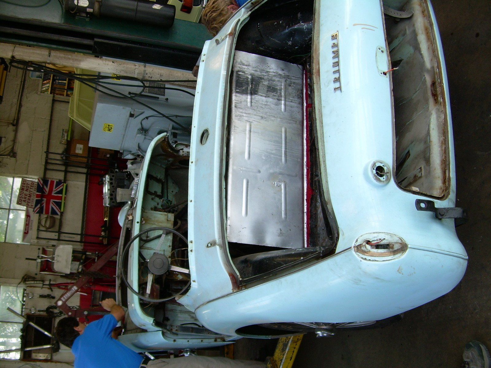 Light blue Triumph car body, partially disassembled, with metal panel. Someone works on the car in a garage.