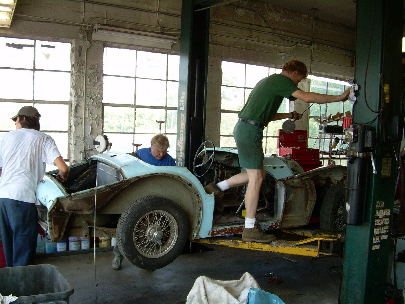 Three people working on a light blue car in a garage with large windows.