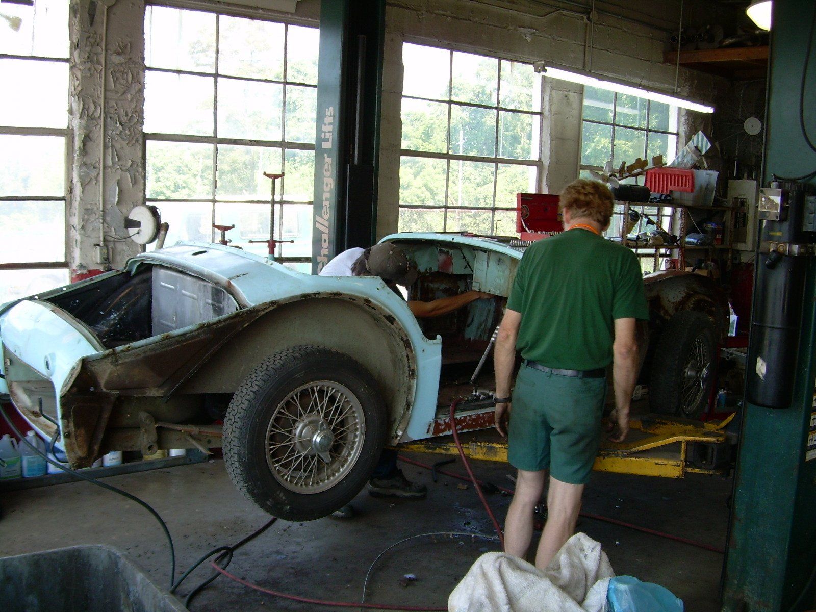 A light blue classic car body being worked on inside a garage; a person in green shorts stands near it.