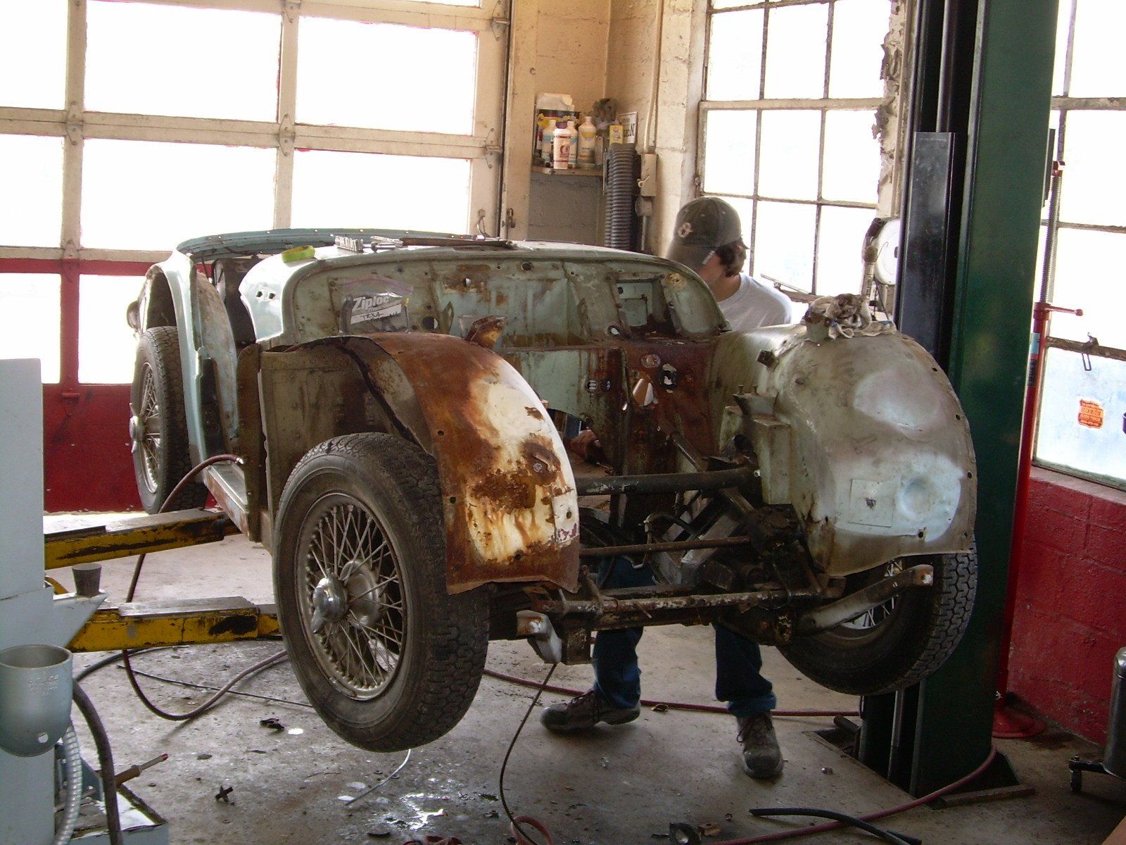 A person works on a rusty vintage car lifted on a hoist in a garage.