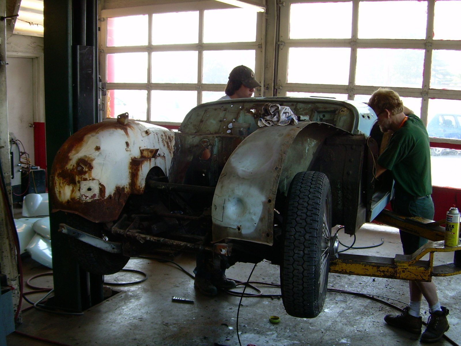 Two people work on a rusty car body lifted in a garage.