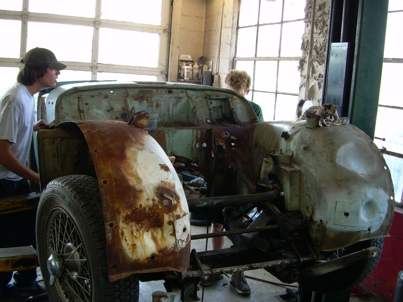 A rusty vintage car stripped down in a garage, two people working on it.