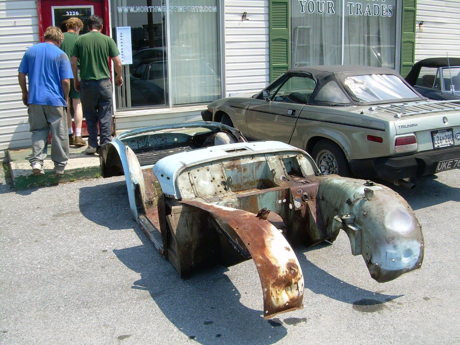Partially disassembled classic car body in front of a shop; two people enter the shop; another car is parked nearby.