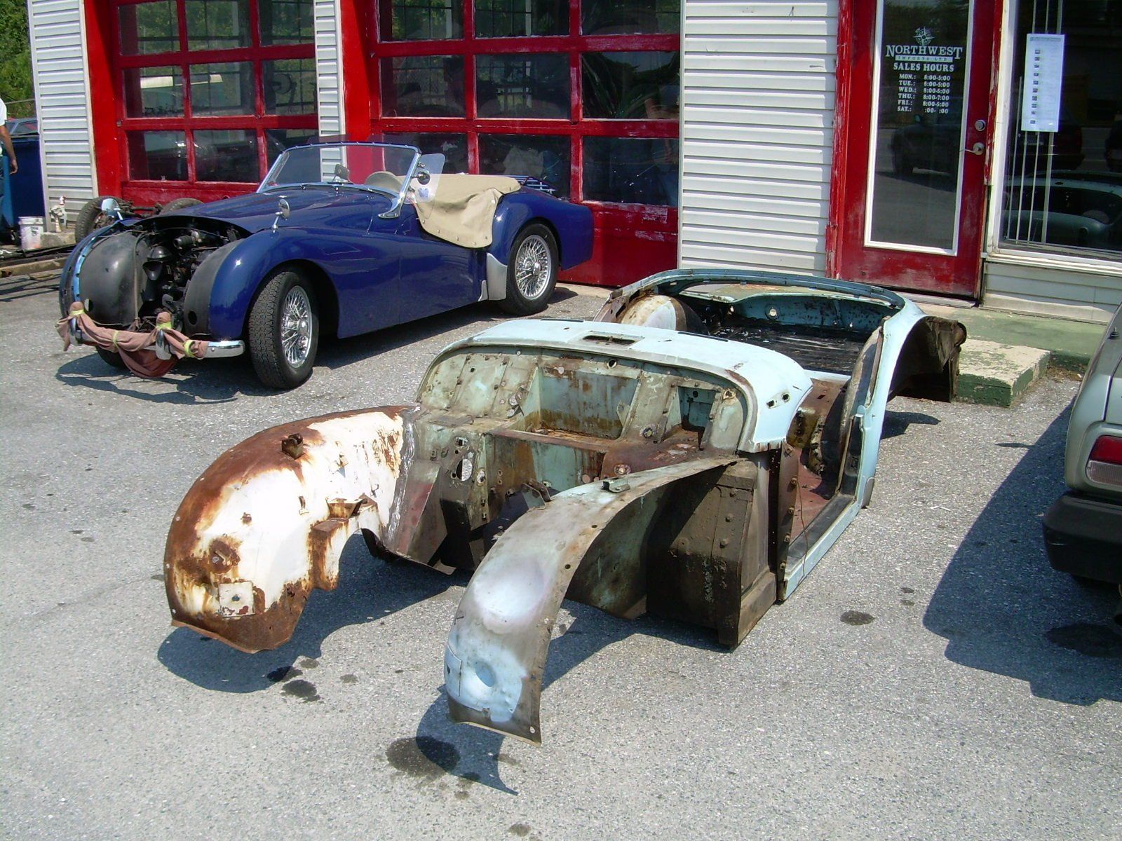 Exterior of a car restoration shop. A disassembled light blue car in foreground; a blue car with beige top in background.