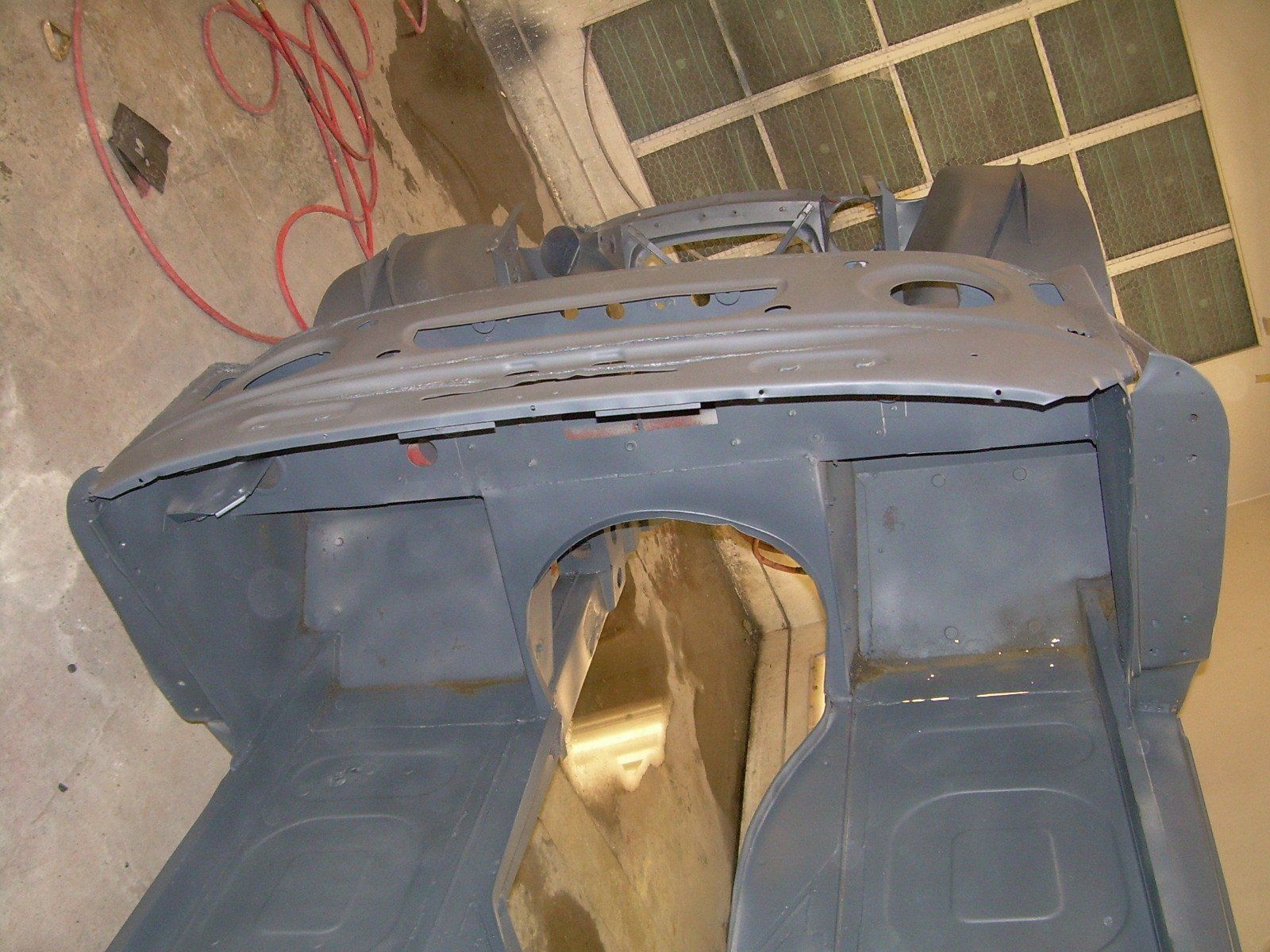 Gray car frame with open interior and engine compartment. Garage setting with tools and wiring.