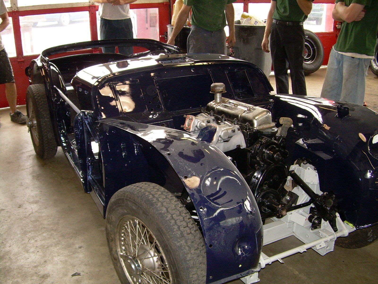 Dark blue vintage car in restoration; engine exposed, wheels on, partially disassembled.