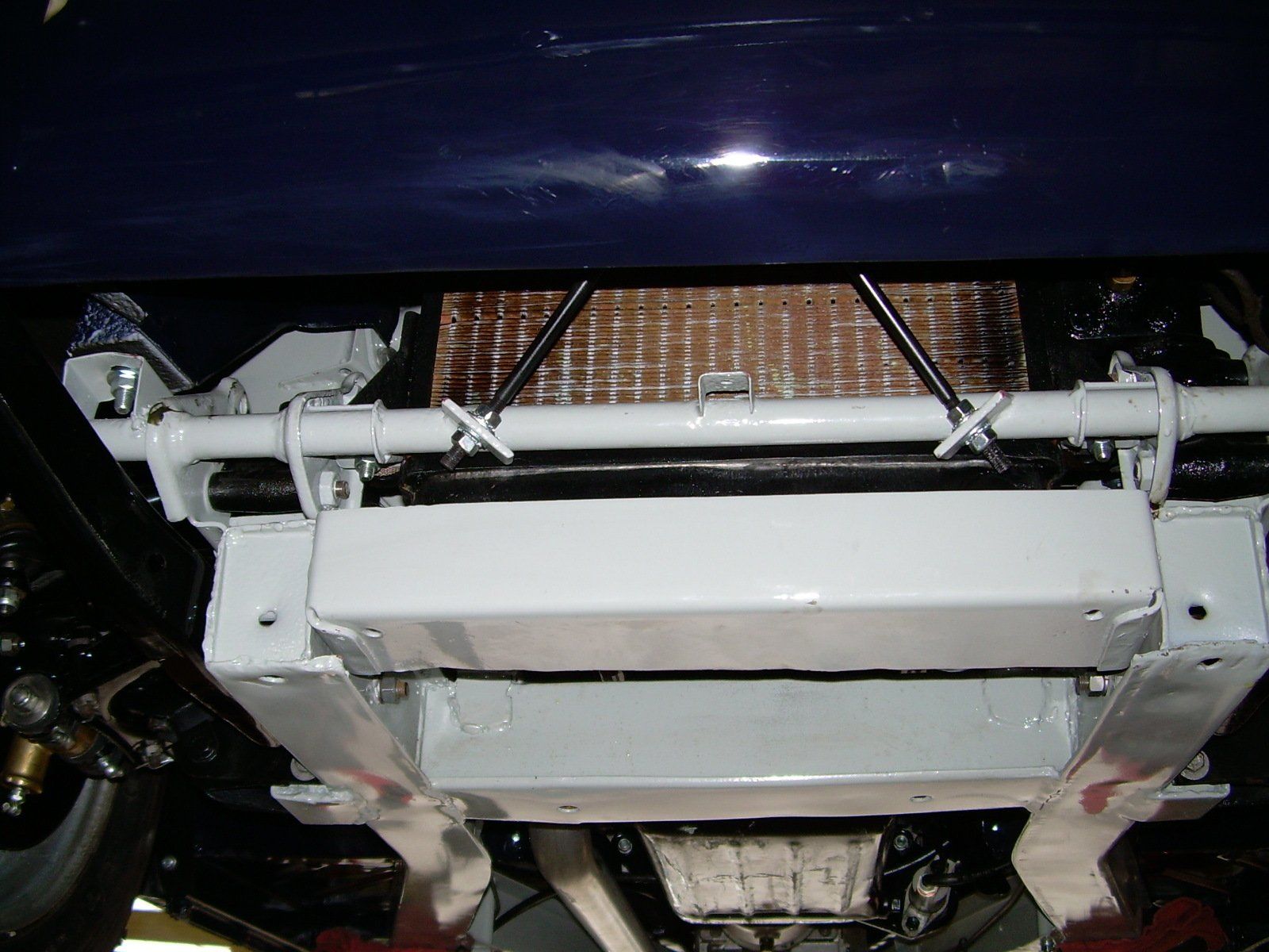 Underside of a light-colored car, showing engine components, radiator, and supportive frame. Dark blue body above.