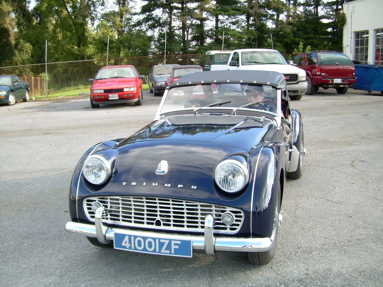 Dark blue Triumph TR3 convertible car parked in a lot, with other vehicles and trees in the background.