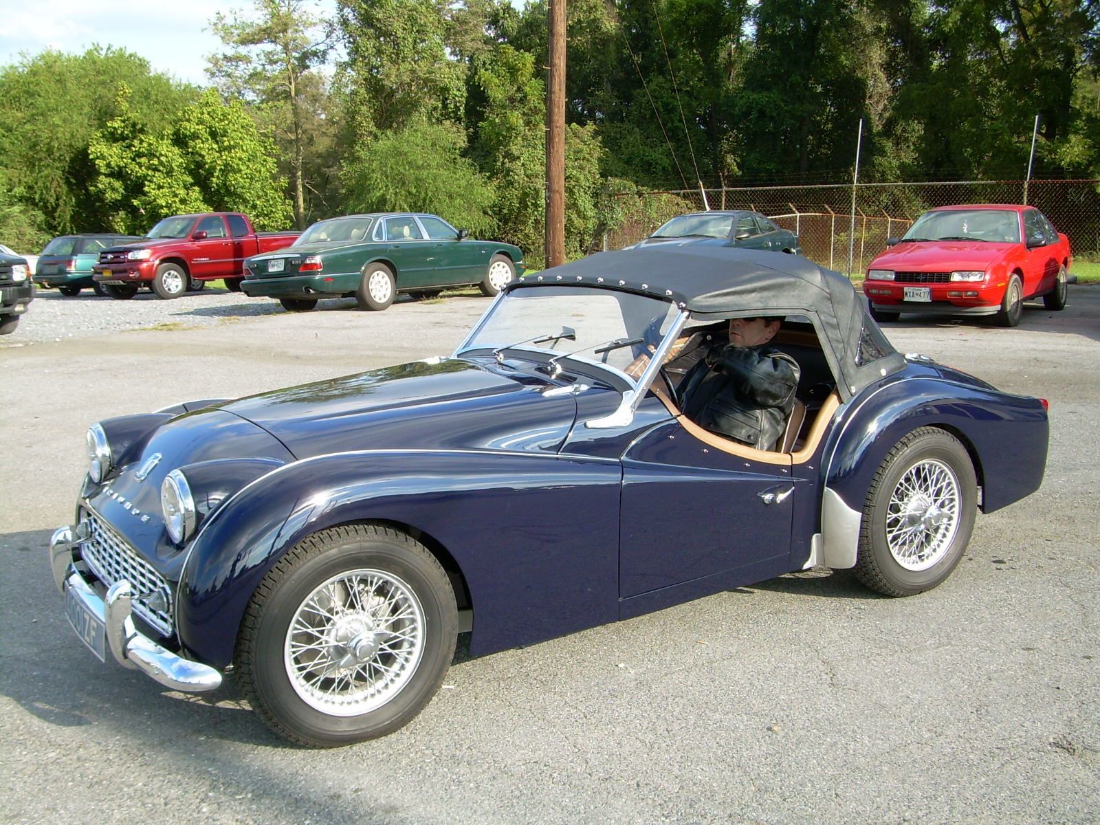 Dark blue Triumph sports car with wire wheels parked in a lot, driver visible.