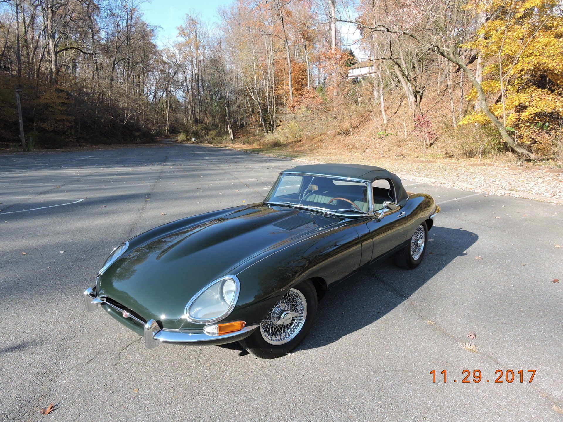 Green Jaguar E-Type convertible on a paved area, with trees in the background under a blue sky.