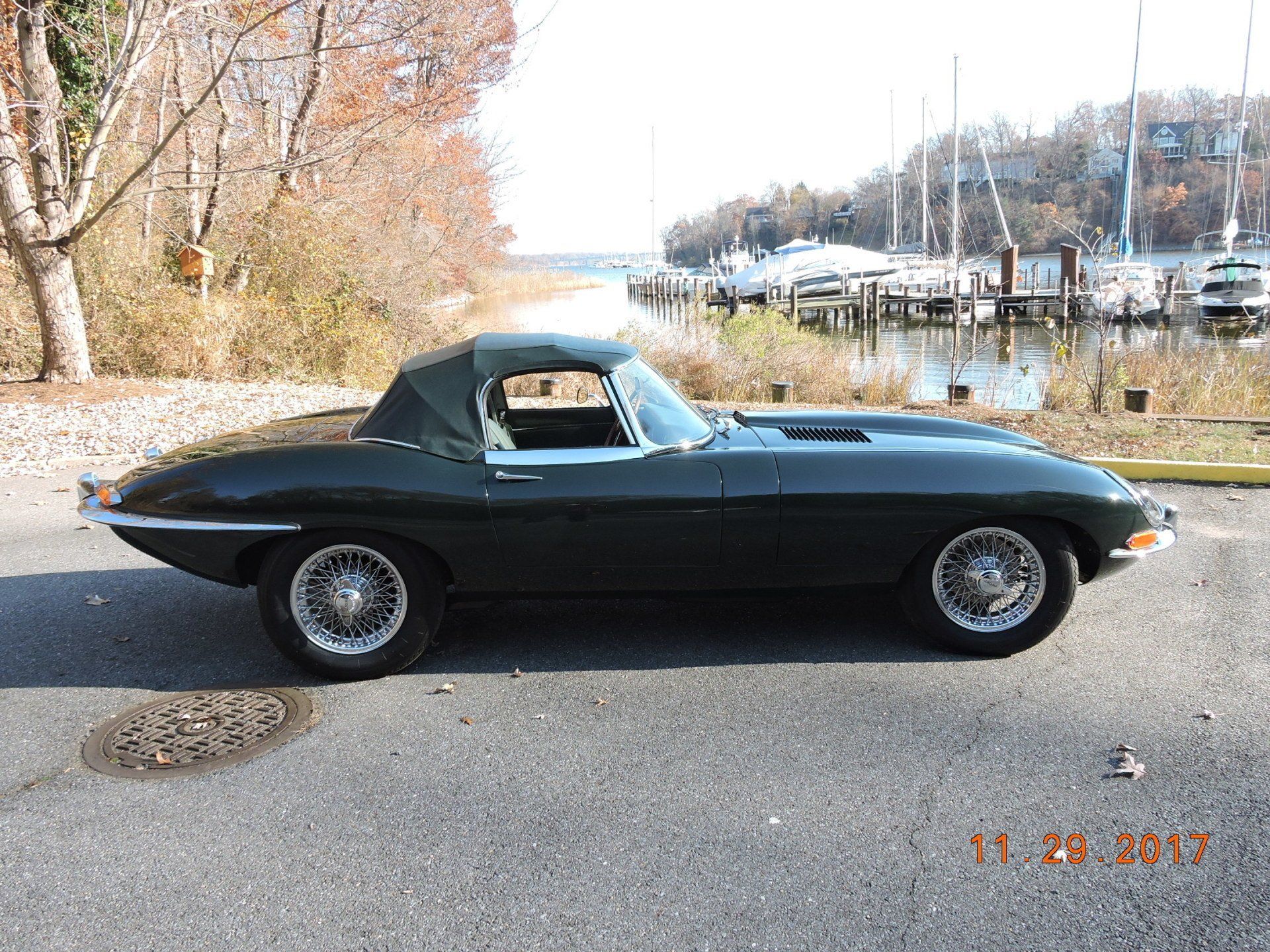 Dark green Jaguar E-Type convertible parked on a paved road near a marina.