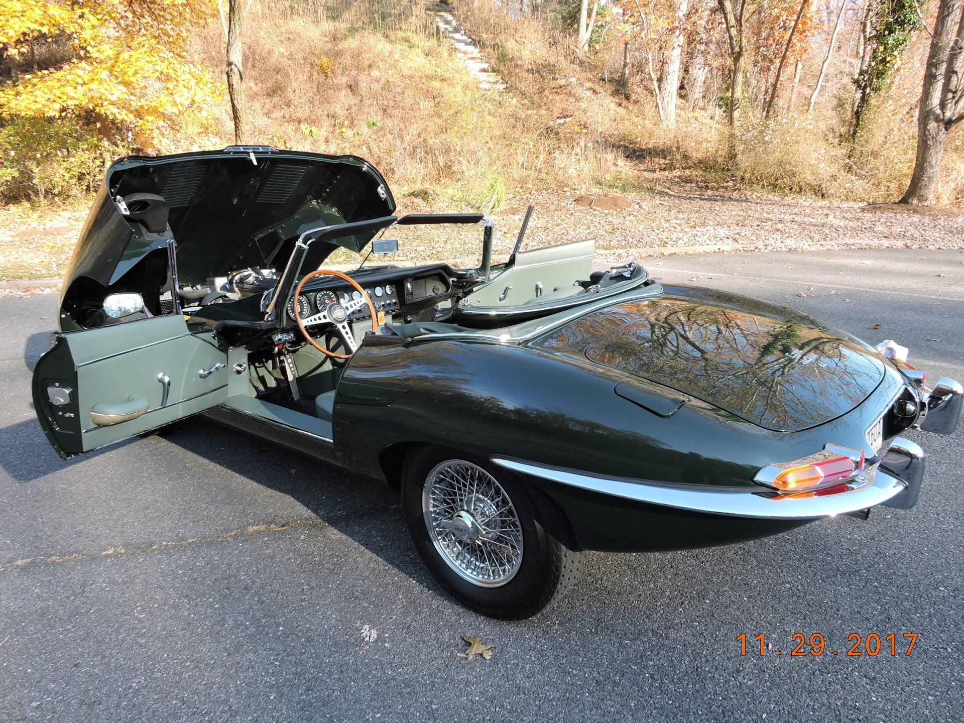 Green Jaguar E-Type convertible with hood and door open, parked on pavement with fall foliage in background.