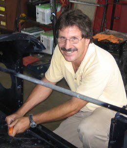 Man working on car frame, smiling. Wearing glasses, tan shirt, and watch. Garage setting.