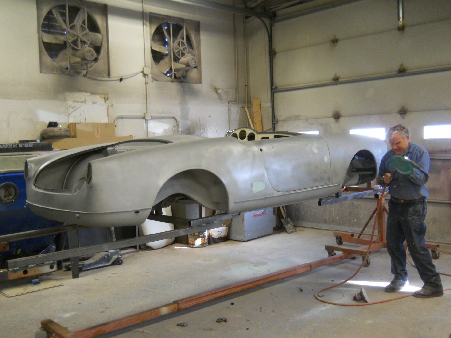 A man working on a silver car body in a workshop. The car is on a metal frame, and air vents are visible.