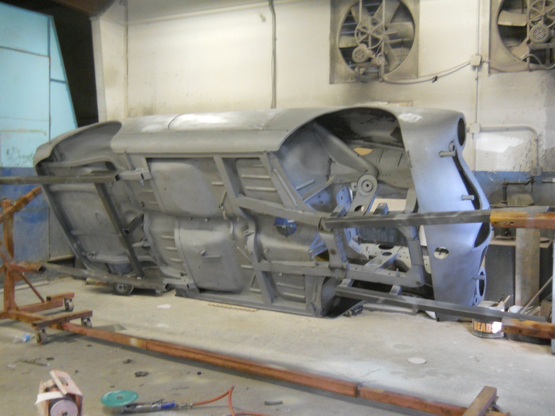 Underside of a car frame in a workshop, grey with metal supports, possibly under restoration.