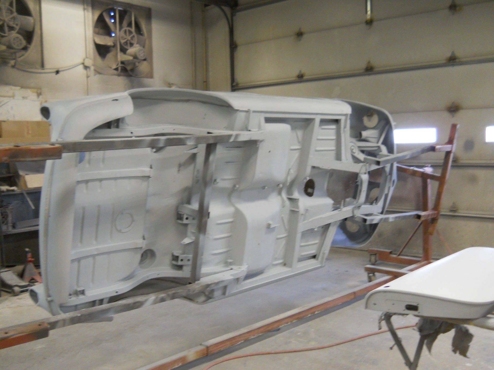 Underside of a classic car chassis, painted white, suspended on a rotisserie in a workshop.