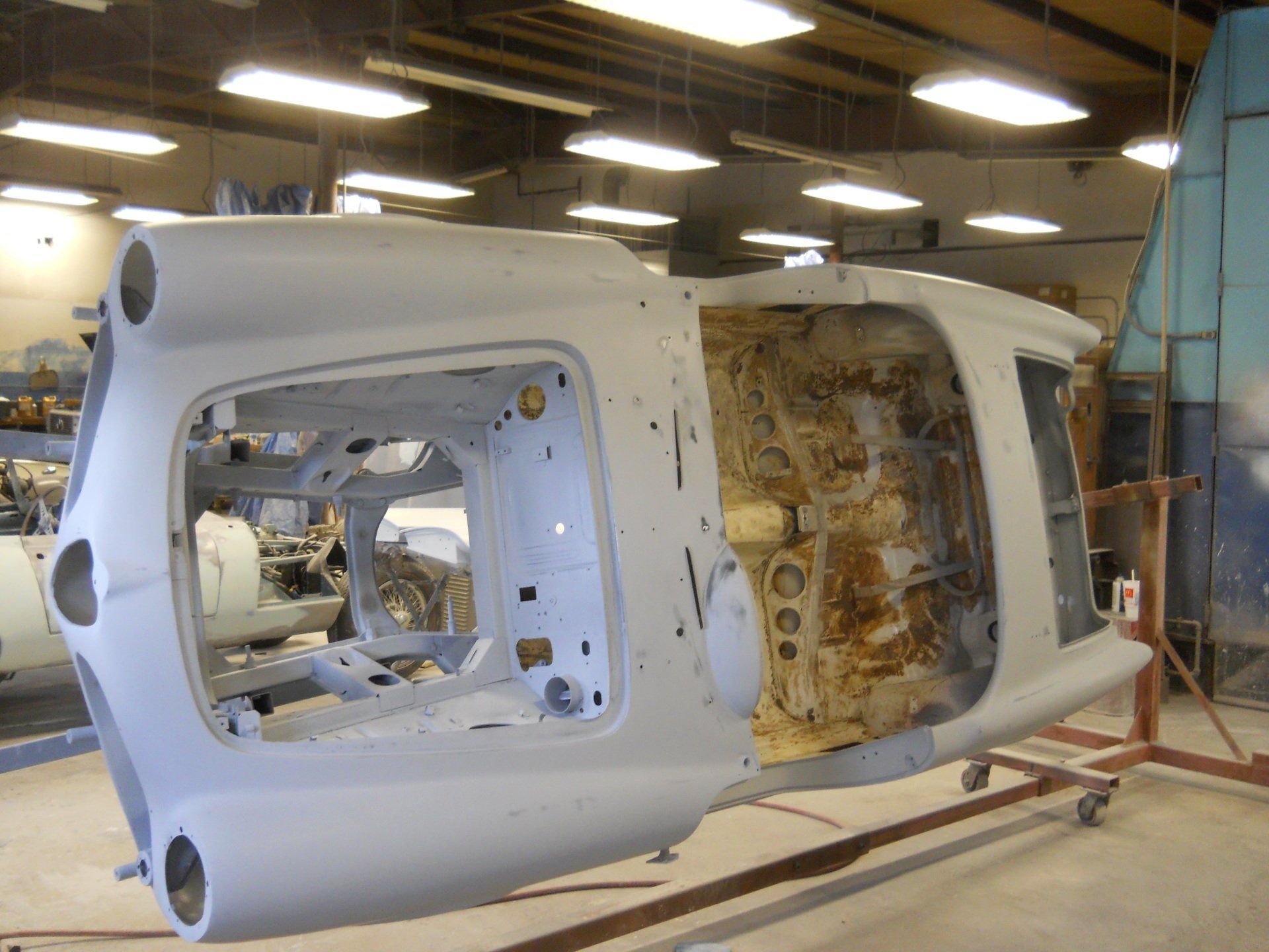 Car body shell undergoing restoration in a workshop; light gray metal on a rolling stand.