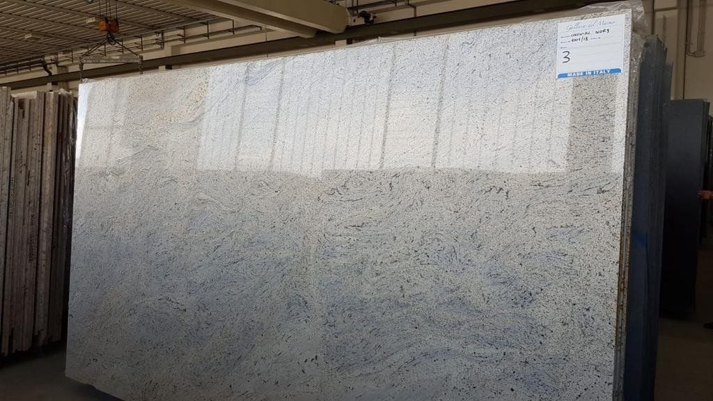 Una grande lastra di granito chiaro, levigata e con venature nere e grigie, esposta in posizione verticale in un magazzino di pietre.