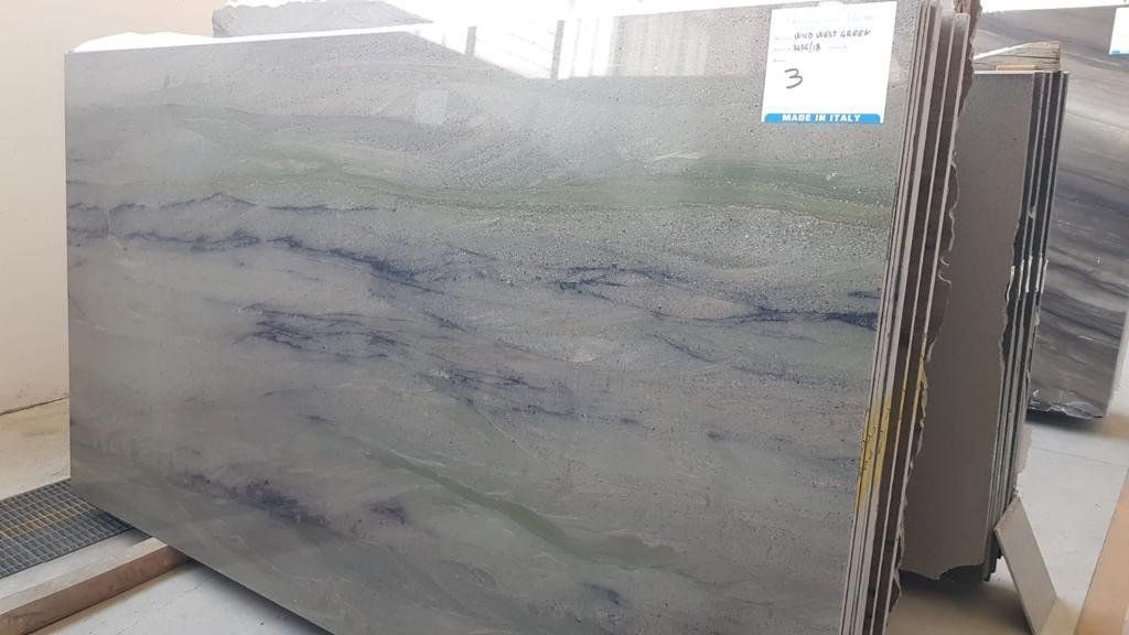 Una grande lastra di granito grigio con venature orizzontali blu e verdi, posizionata verticalmente in un magazzino.