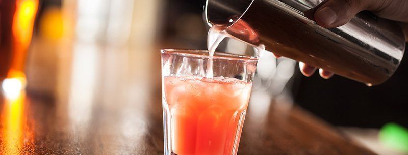 A hand pours a pink cocktail into a clear glass at a bar, with warm blurred lights in the background.