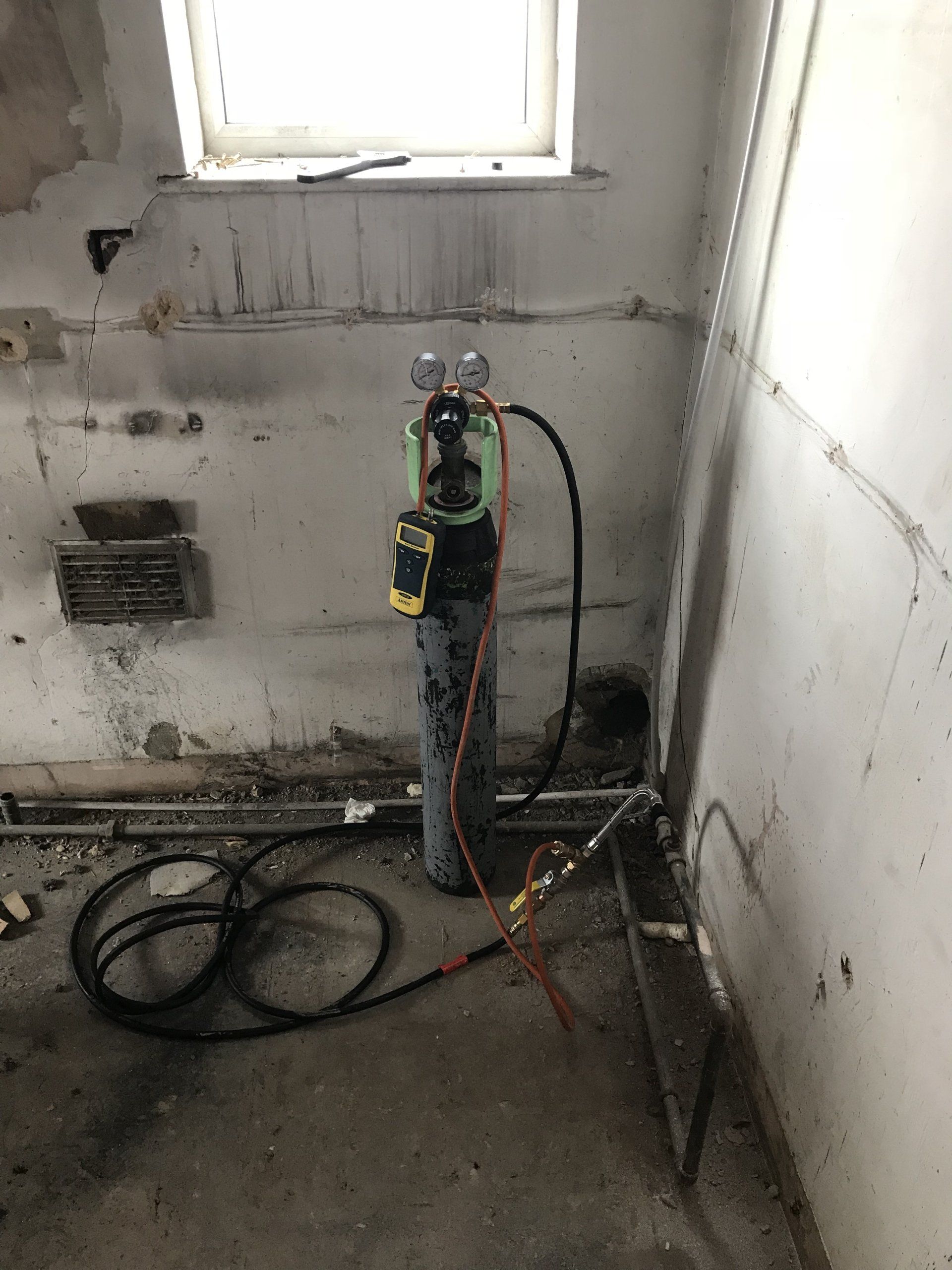 An oxygen tank with gauges in a construction site. Hoses run along the floor. Grimy, damaged walls and window.