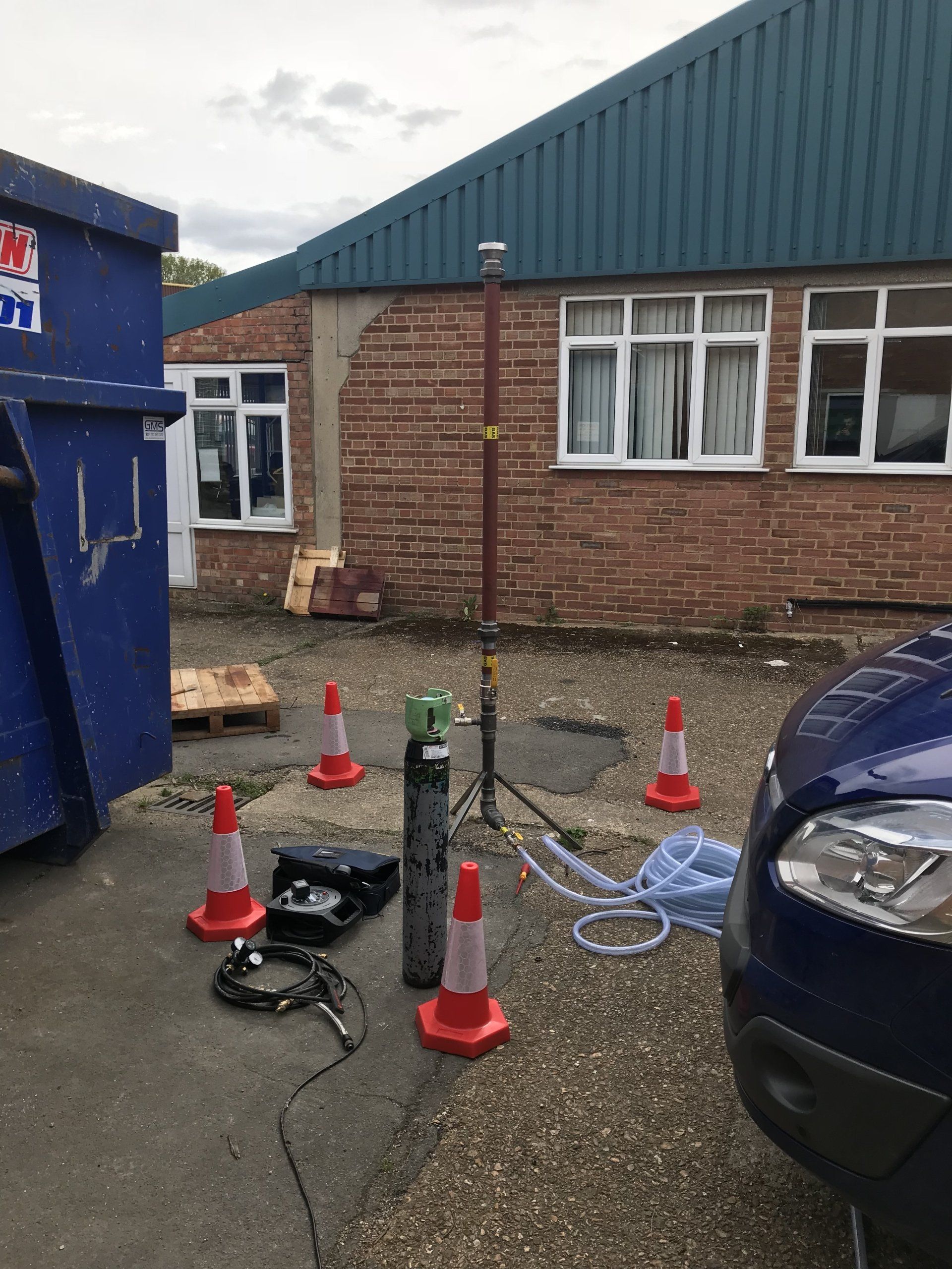Outside a brick building, equipment and cones surround a car, possibly surveying or testing.