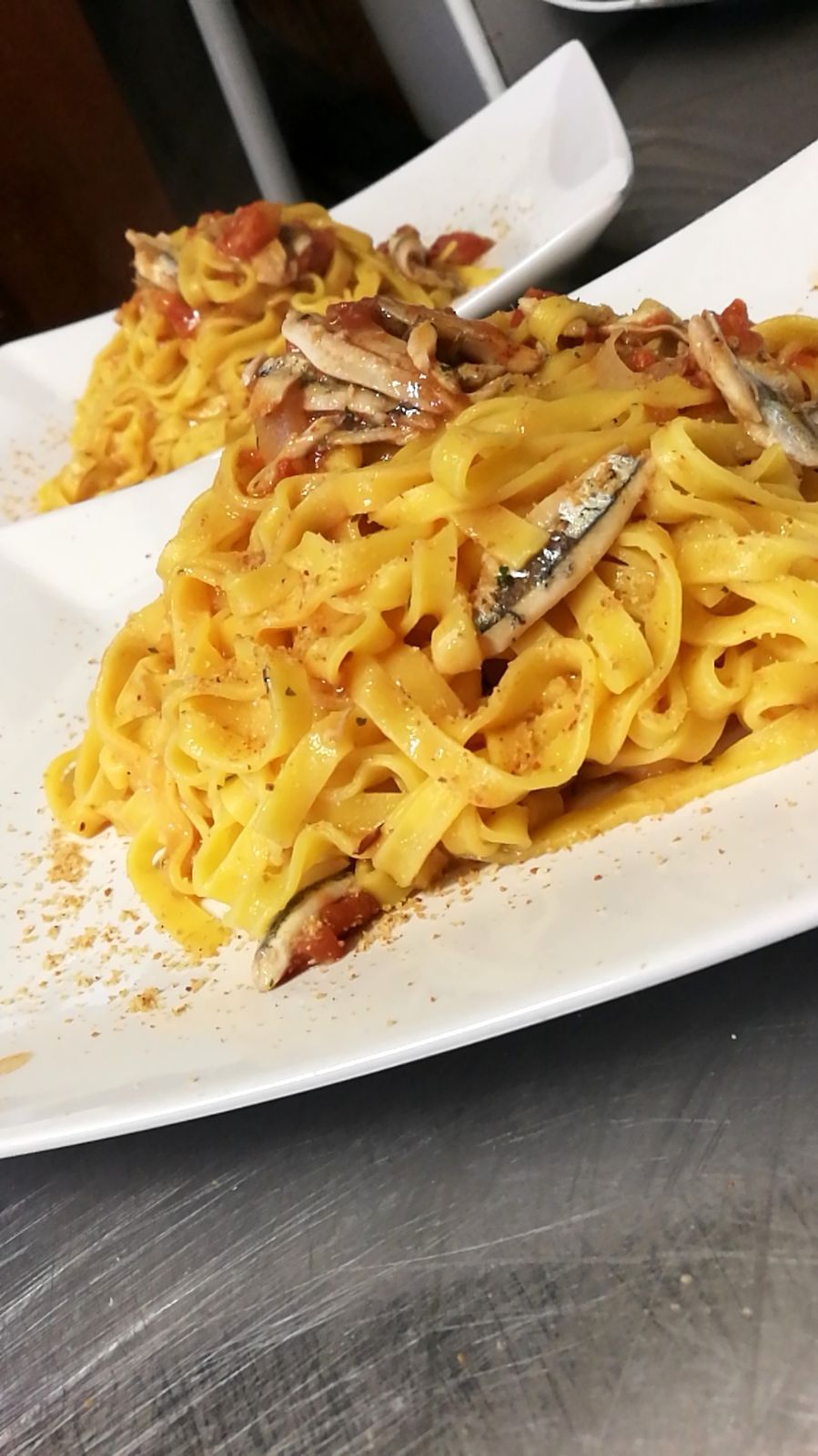 Due piatti di pasta con sugo e contorno di pesce su una superficie in acciaio inox.