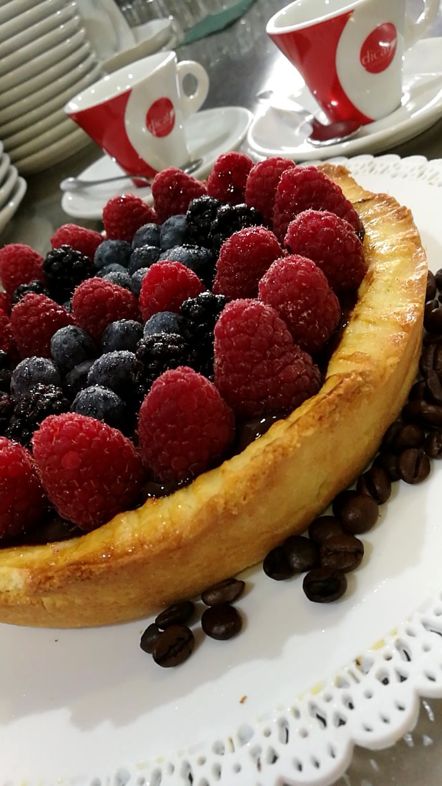 Crostata di frutti di bosco con lamponi, mirtilli, more e chicchi di caffè su un centrino; sullo sfondo tazzine da caffè.