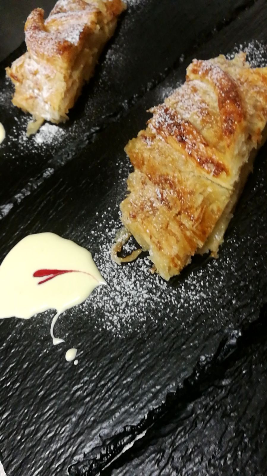 Strudel di mele servito su piatti di ardesia nera, condito con panna e spolverato con zucchero a velo.
