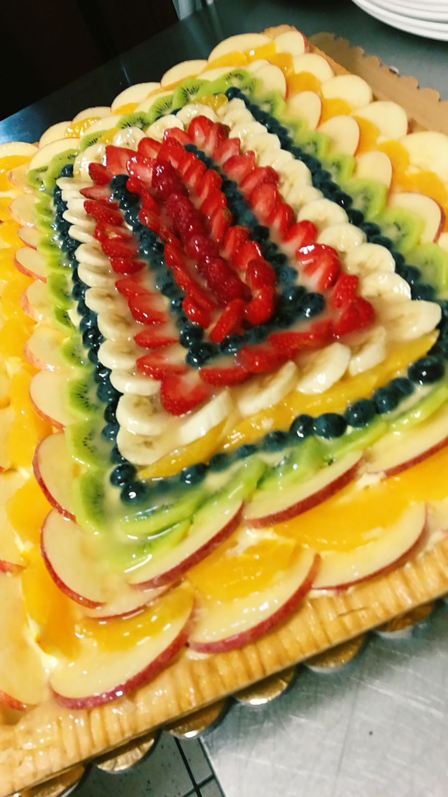 Crostata di frutta quadrata con anelli concentrici di vari frutti, guarnita con fragole rosse.