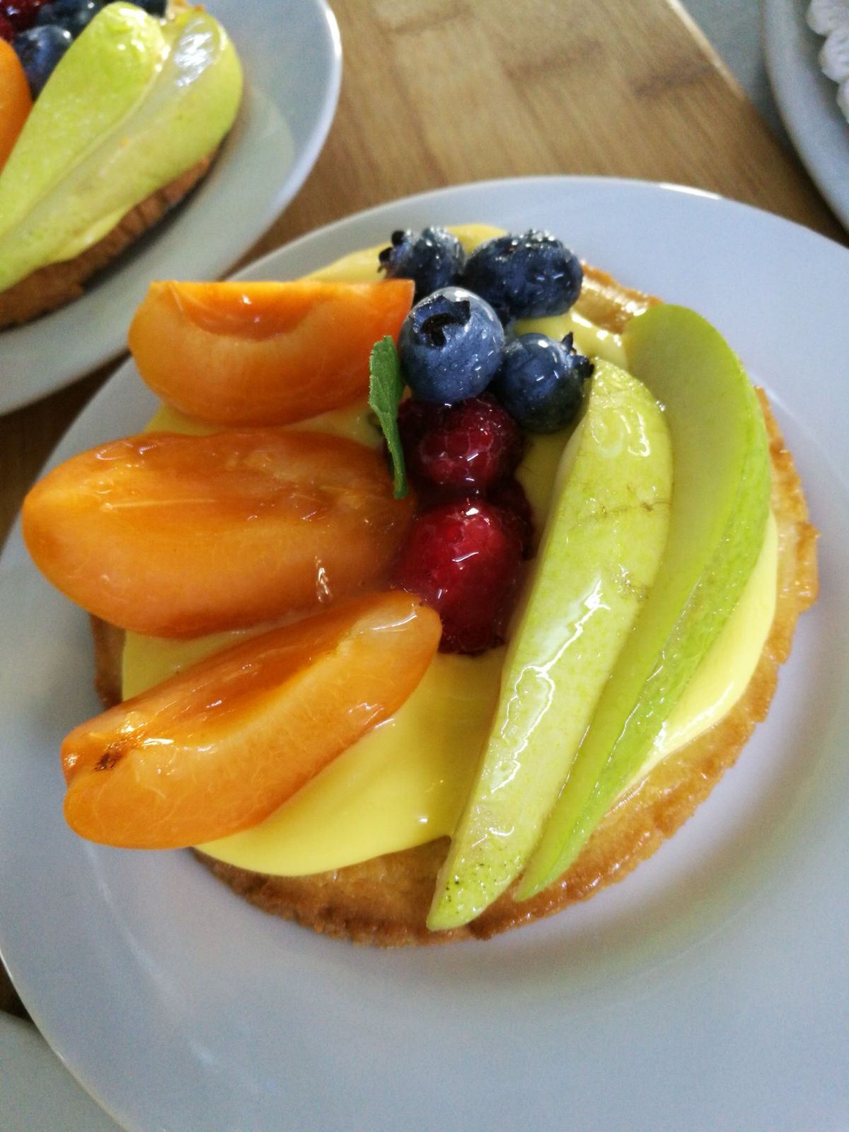 Crostata di frutta con crema pasticcera gialla, albicocche, mirtilli, lamponi e fette di pera su un piatto bianco.