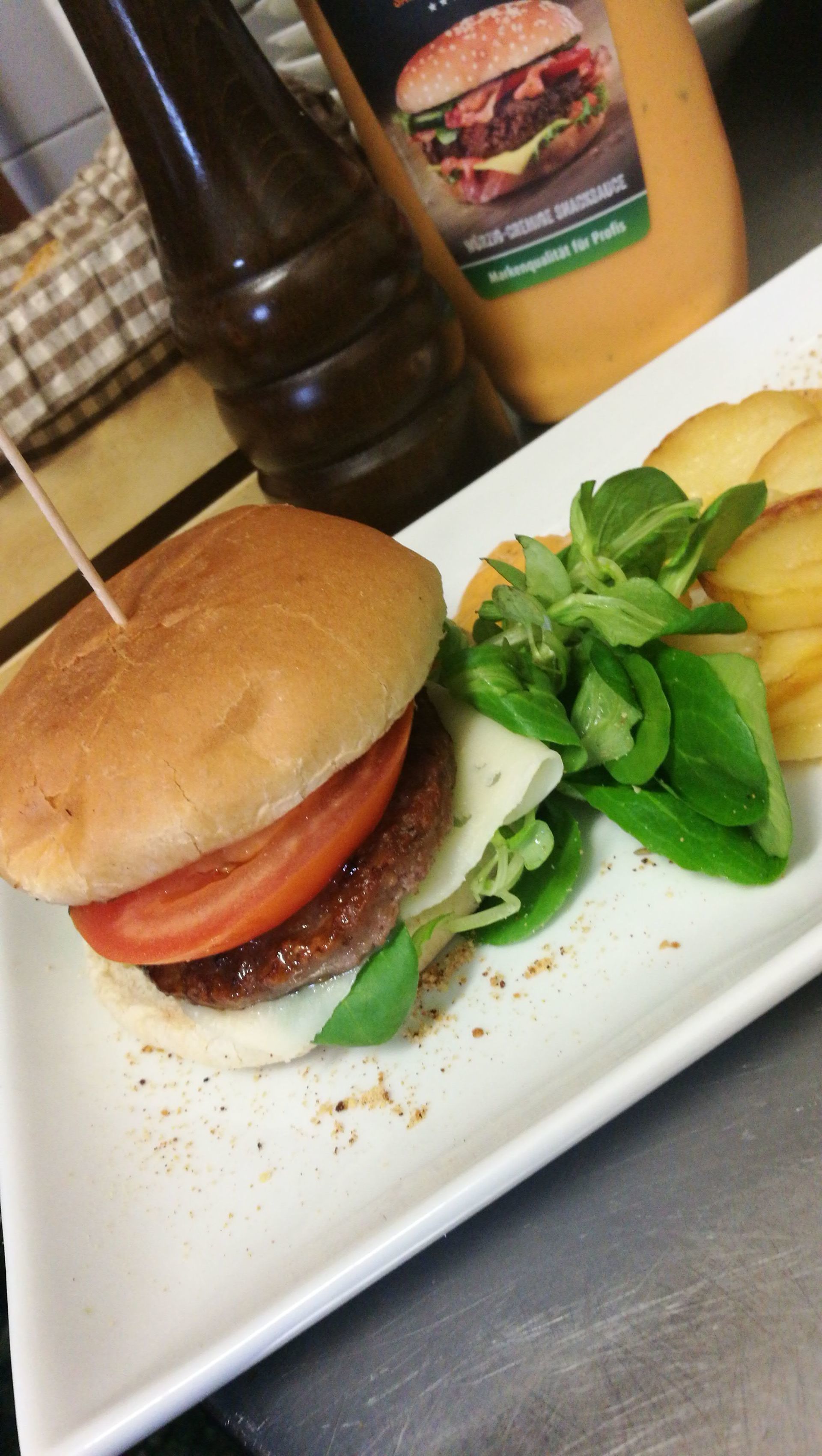 Hamburger su un piatto con patatine fritte e verdure, accanto a salsa e una bottiglia.