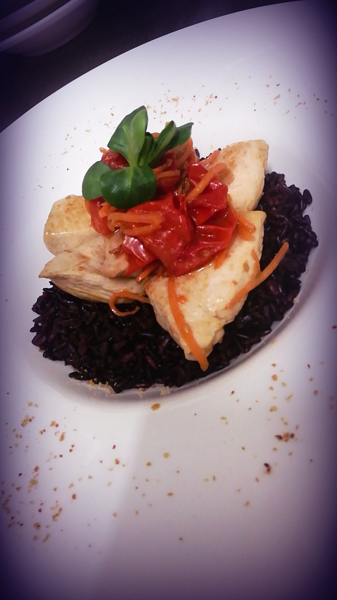 Piatto di pollo e pomodoro su riso nero, guarnito con verdure, servito su un piatto bianco.