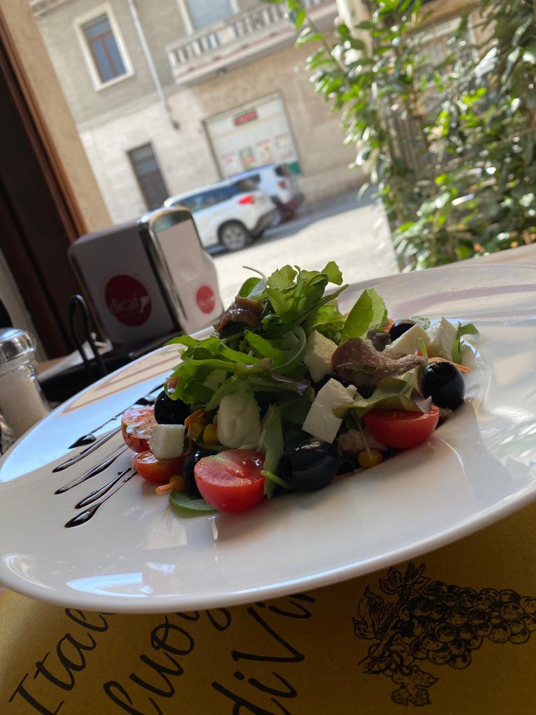 Insalata su un piatto bianco con pomodori, olive, rucola e formaggio, vicino a una finestra che si affaccia su una strada.