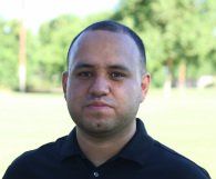 A man in a black shirt is standing in a field and looking at the camera.