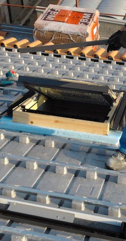 Un operaio installa il telaio di una finestra per tetti su un tetto ricoperto da pannelli isolanti termici grigi e file di tegole in ceramica.