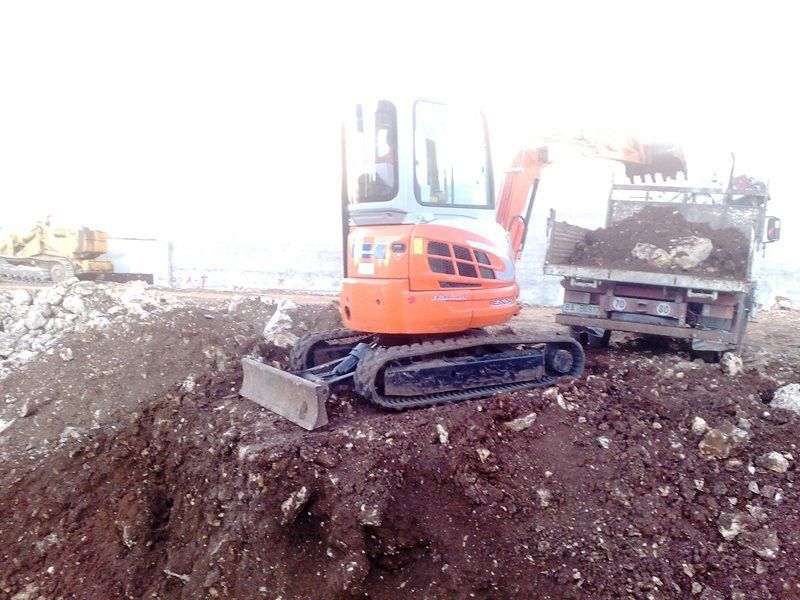 Un mini-escavatore arancione carica terra marrone in un autocarro ribaltabile in un cantiere.