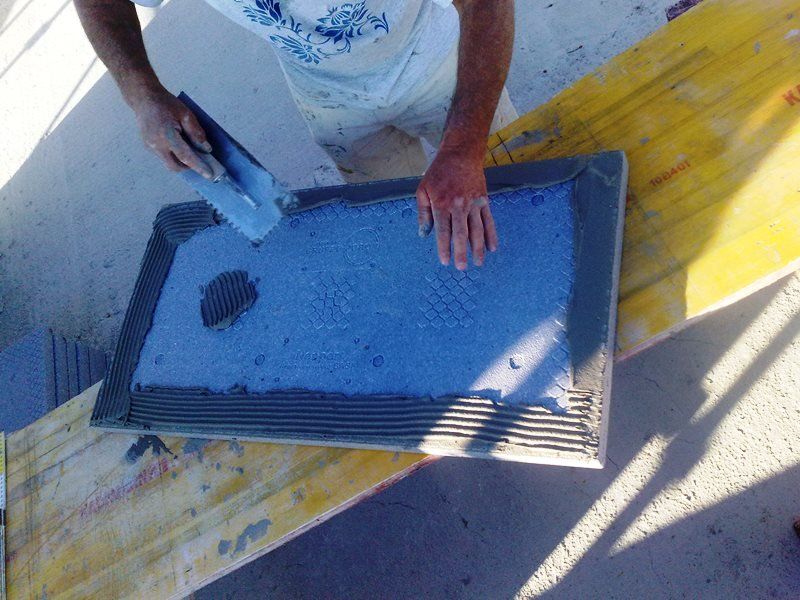 Una persona utilizza una spatola dentata per stendere la colla grigia per piastrelle sul retro di una piastrella blu.