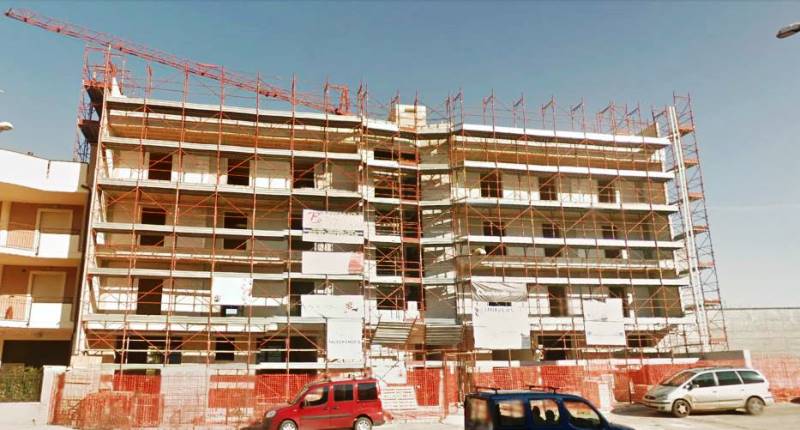Un edificio residenziale a più piani in costruzione, ricoperto di impalcature e con cartelloni pubblicitari sulla facciata.