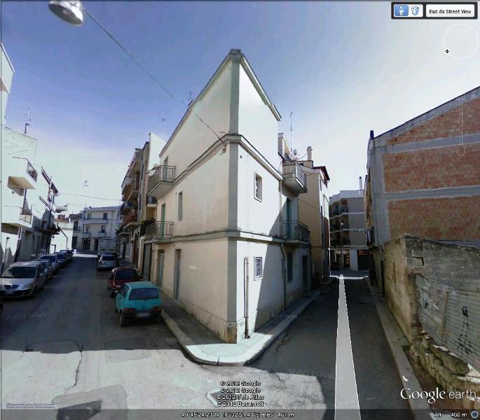 In una cittadina, all'incrocio di una strada, si erge un edificio bianco dagli angoli acuti, con auto parcheggiate sulla sinistra e un muro di pietra.