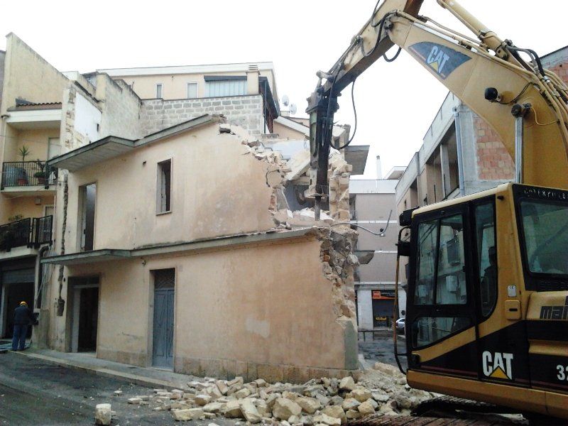Un escavatore Caterpillar giallo sta demolendo l'angolo superiore di un edificio beige a due piani in una stretta via cittadina.