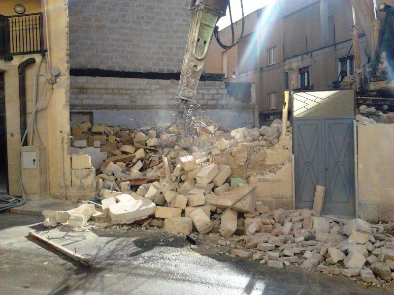 Un escavatore con martello idraulico frantuma un cumulo di macerie di pietra tra due edifici urbani.