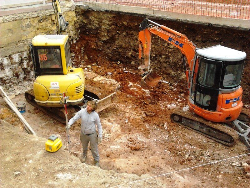 Una persona si trova in uno scavo edile tra un miniescavatore giallo e uno arancione.