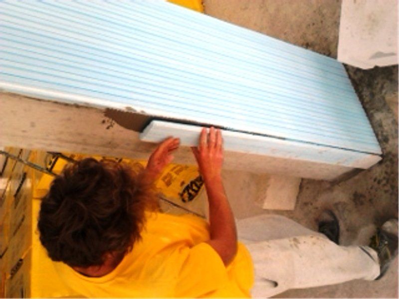 Una persona con una maglietta gialla installa un pannello isolante blu su una parete di cemento utilizzando della colla.