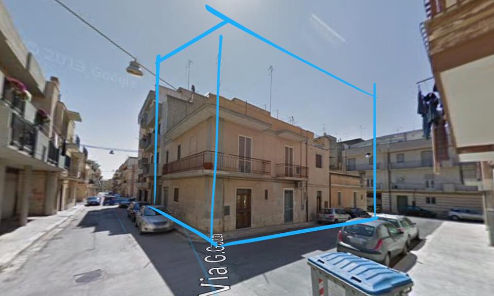 Un edificio beige a due piani all'angolo di una strada, evidenziato da linee blu, con auto parcheggiate nelle vicinanze sotto un cielo azzurro e limpido.