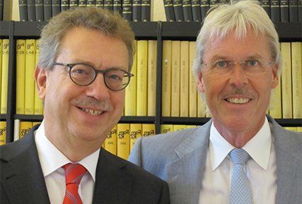 Zwei Männer in Anzug und Krawatte posieren vor einem Bücherregal für ein Foto.